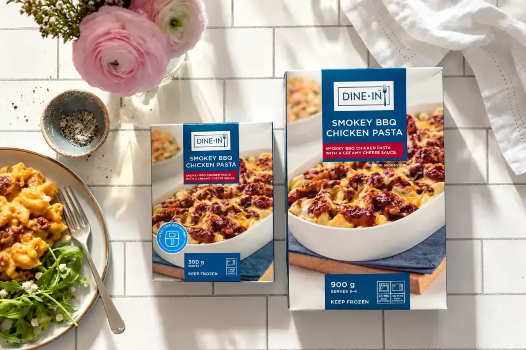 DINE-IN Smokey BBQ Chicken Pasta frozen meals in 300 g and 900 g packs, displayed on a sunlit tiled table.